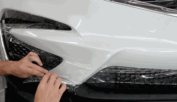 A pair of hands smooth out paint protection film across the front bumper of a white SUV, working carefully around the headlights and grille.