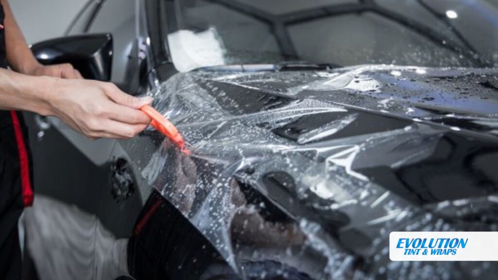 A technician uses a red tool to install paint protection film on the hood of a black car, with soapy water aiding the smooth application.