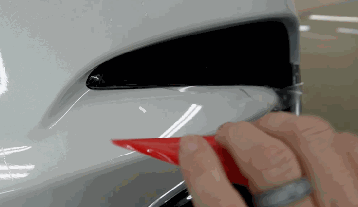 A close-up of a hand using a red squeegee to press paint protection film onto the edge of a white car’s headlight area, ensuring a seamless seal.